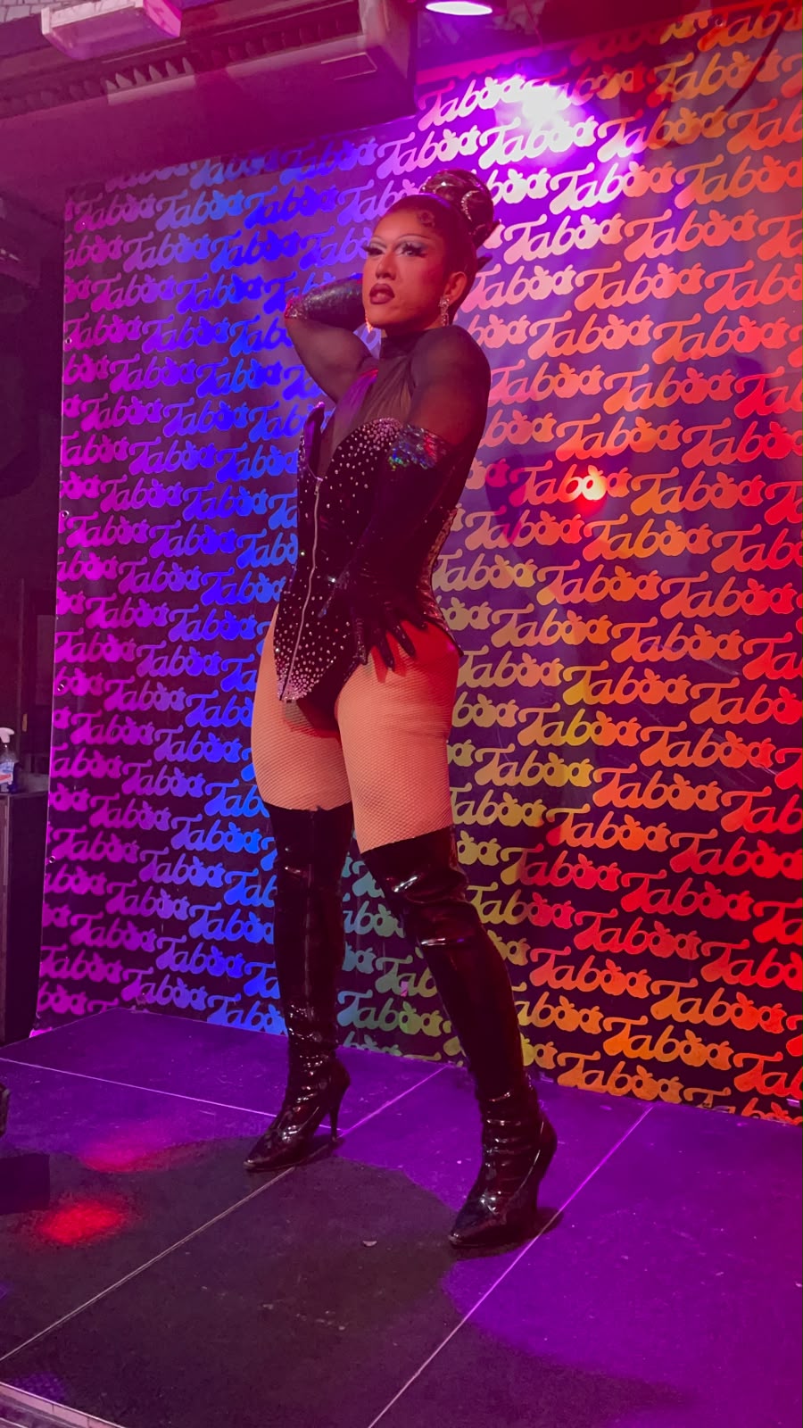 Drag performer on stage in front of a wall covered in the Taboo bar logo