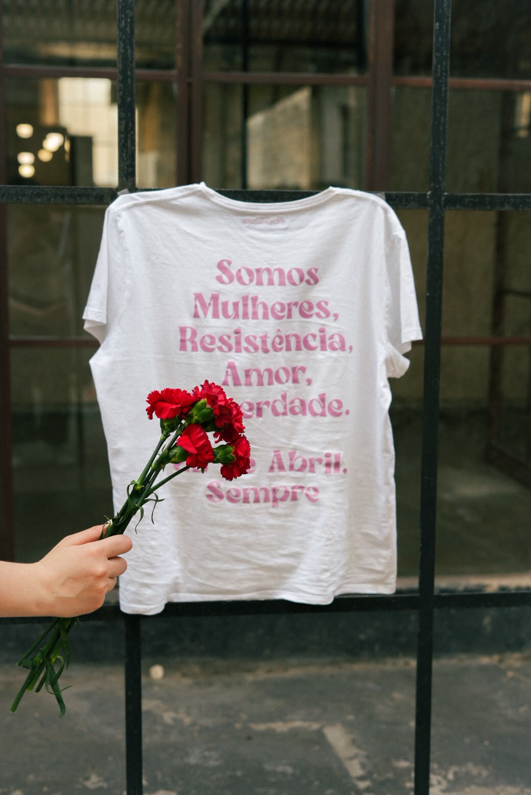 White t-shirt printed 'Somos Mulheres, Resistência, Amor, Liberdade. 25 de Abril, Sempre' with red carnations