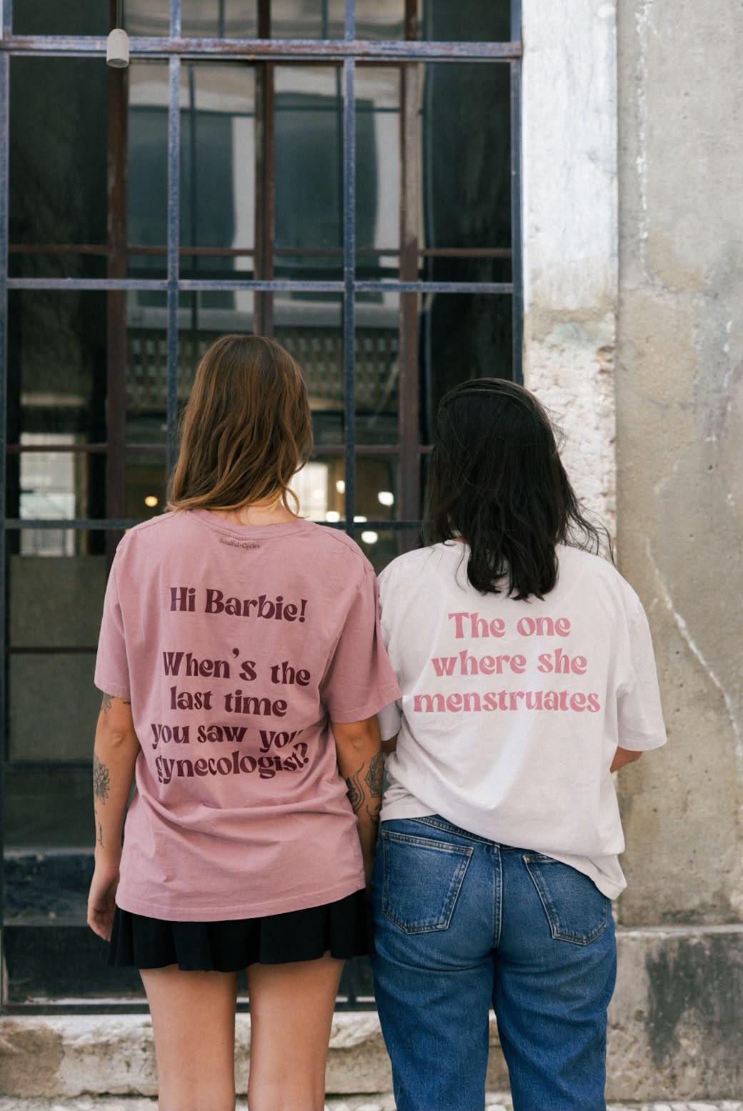 Two people from behind wearing t-shirts printed 'Hi Barbie! When's the last time you saw your gynecologist?' and 'The one where she menstruates'