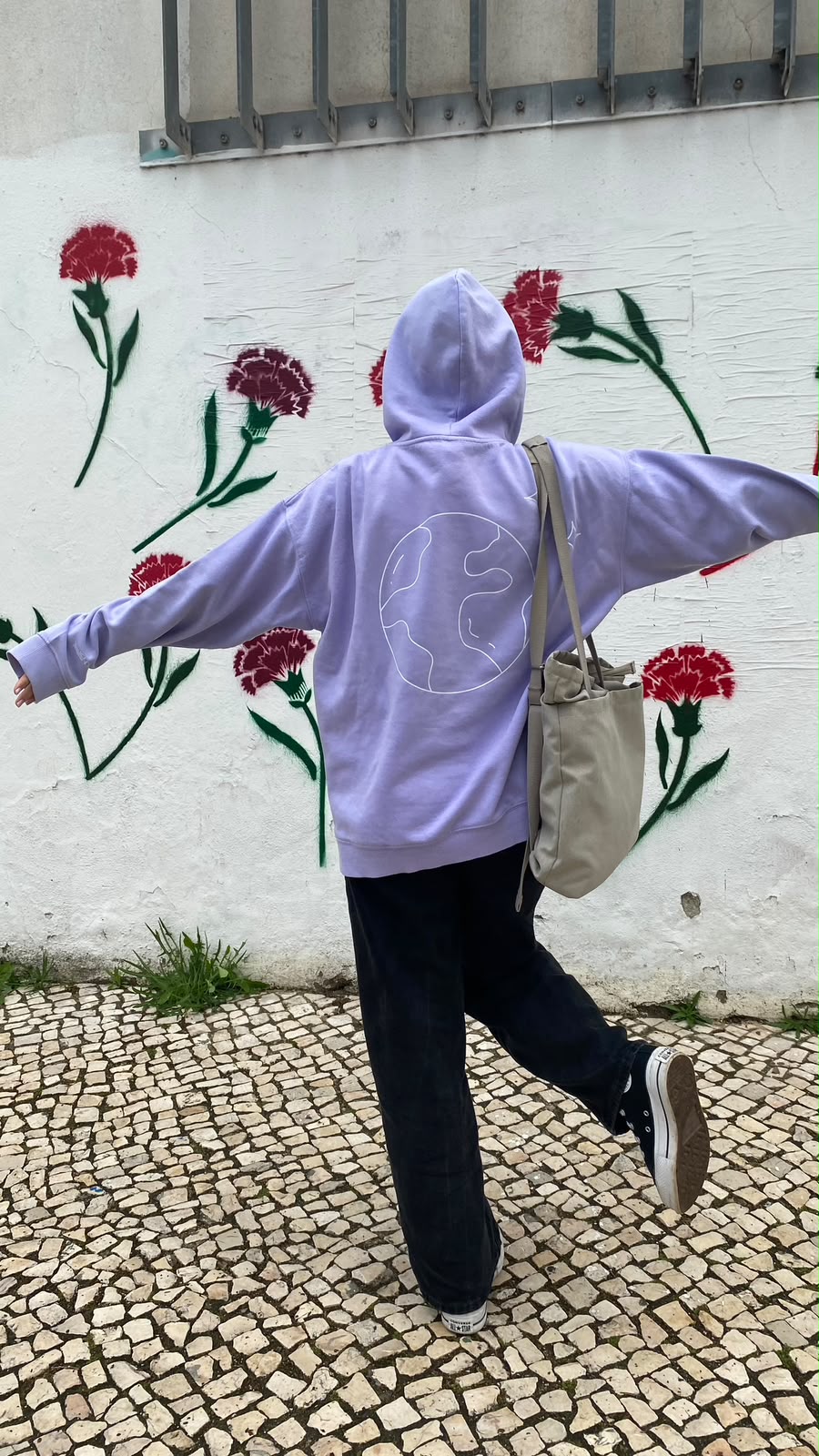Person in a lilac hoodie with a globe drawing, dancing in front of a wall painted with red carnations