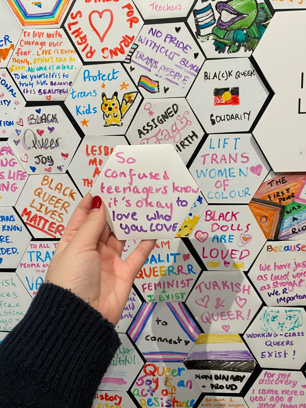 Hand holding a hexagon tile reading 'So confused teenagers know it's okay to love who you love' in front of a wall of queer messages